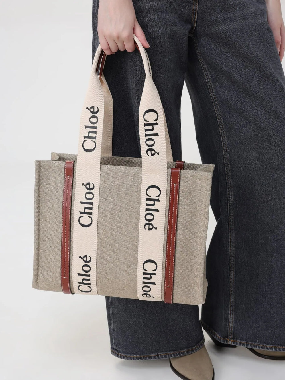 A person wearing dark wide-leg jeans holds a beige canvas tote bag with cream-colored shoulder straps printed with the name 'Chloé' in black. The bag has brown leather trim and a brown zipper.
