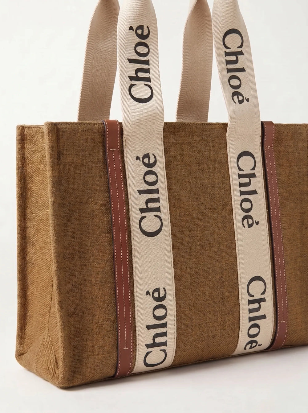 A brown woven tote bag with cream-colored shoulder straps featuring the black printed brand name 'Chloé' repeatedly, bordered by thin reddish-brown stripes.