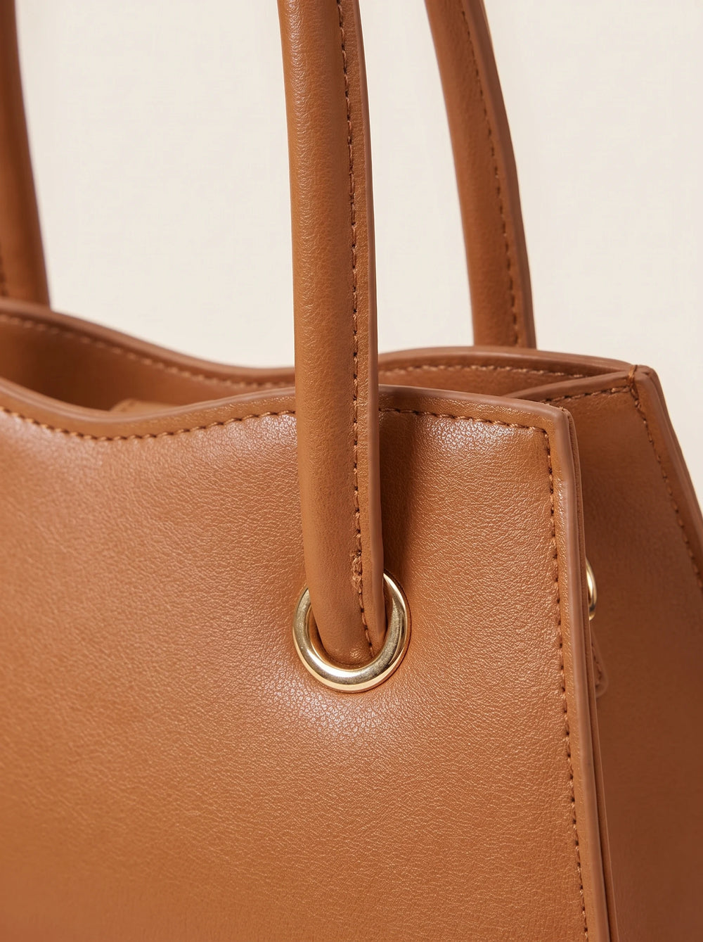 Close-up of a tan leather handbag showing the side profile, double top handles, and a gold-tone metal grommet detail against a light beige background.