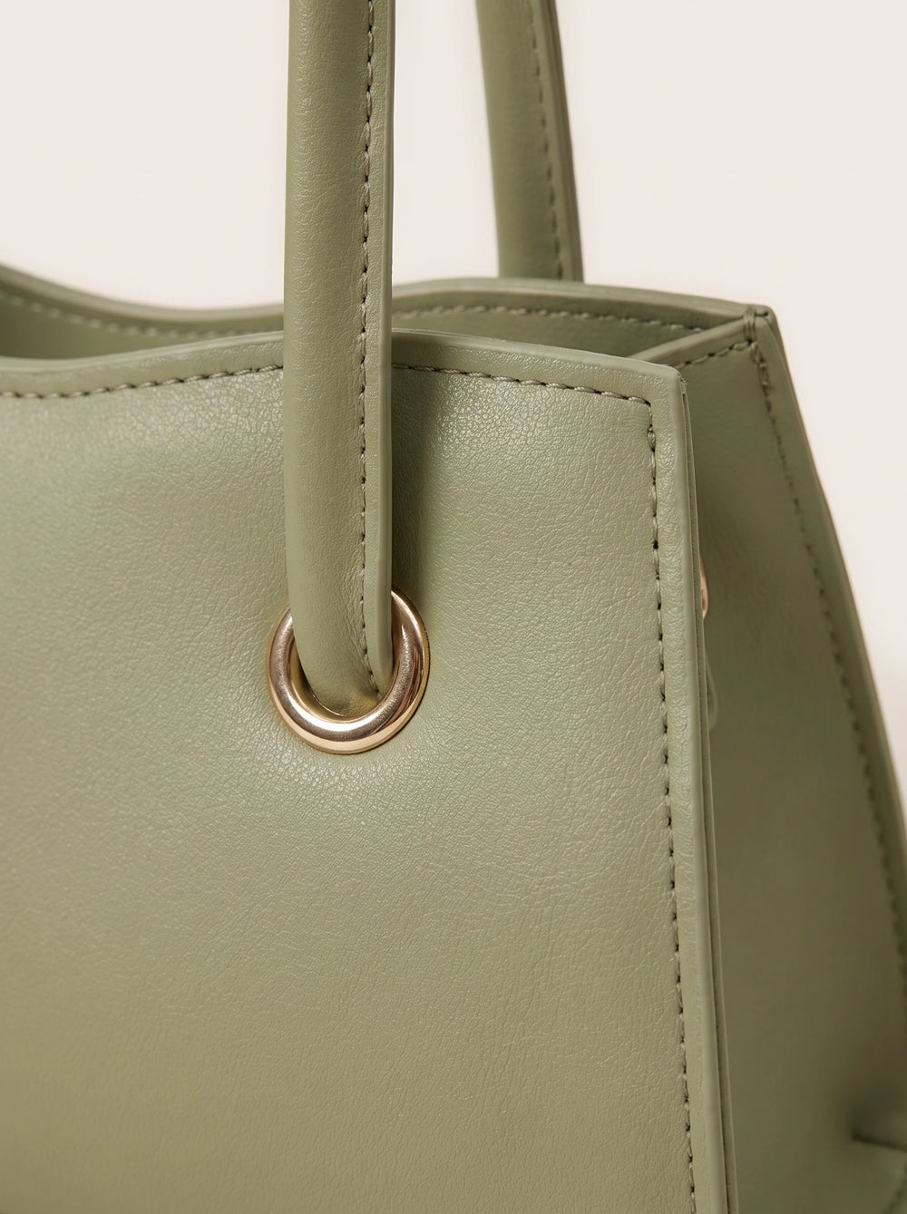 Close-up of a structured tote bag in sage green leather with a rose gold metal grommet and handle attachment, set against a plain off-white background.